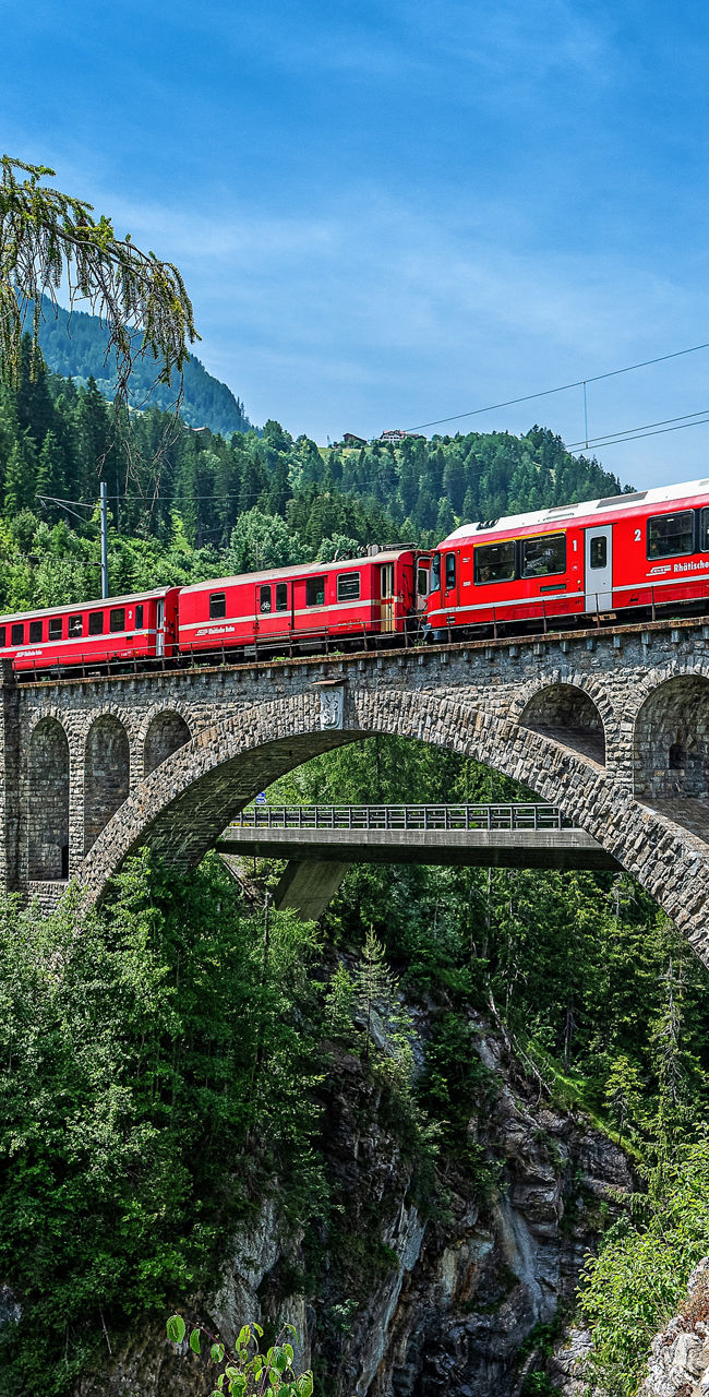 Red train crossing stone bridge in mountains
